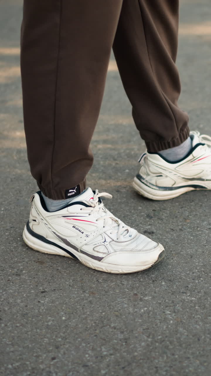 zapatillas blancas sobre asfalto, paso informal, pantalones de chándal marrones remangados por encima de los tobillos, deportivas desgastadas con cordones visibles y suelas texturizadas, primer plano de pies en movimiento sobre pavimento iluminado por el sol, trayecto tranquilo