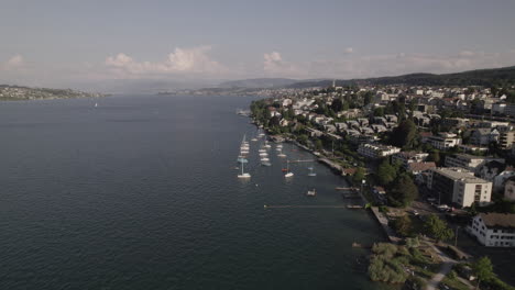 Drone-shot-flying-near-the-city-of-Thalwil-at-the-Zurich-Lake-in-Switzerland-on-a-sunny-day-with-water-and-boats-around-the-shore-and-white-houses-in-the-city-LOG