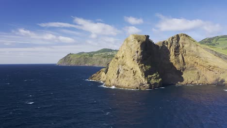 dramatic-vulcanic-cliffs-revealing-Rosais-village,-green-vegetation-and-lava-formations-next-to-the-sea-drone-footage,-Sao-Jorge-island,-Azores,-Portugal