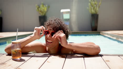 Un-Joven-Birracial-Se-Relaja-Junto-A-La-Piscina-En-Casa,-Gafas-De-Sol-En-La-Mano