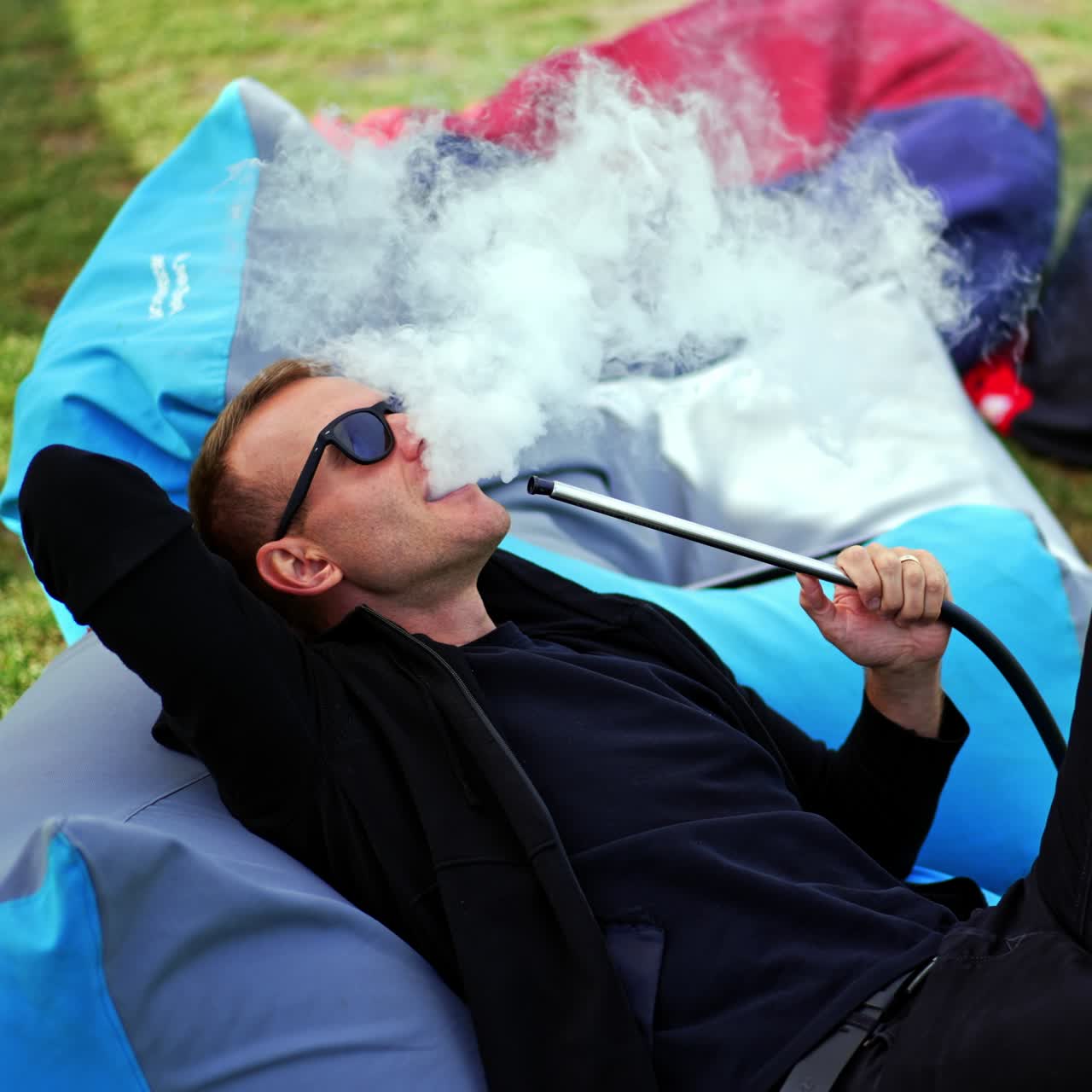 Relaxed man in black clothes and sunglasses lies in bean bag chair outdoors. Resting man smoking hookah producing smoke