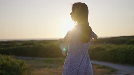 Beautiful-dark-haired-woman-wearing-dress-swings-arms-around-at-sunset-by-beach