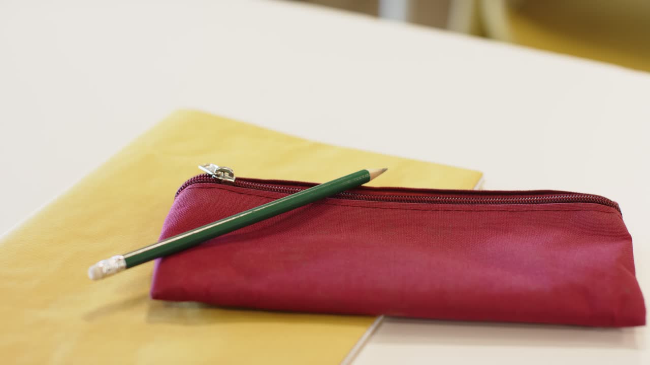A câmera está mudando o foco na mesa branca para enfatizar o estojo vermelho com zíper e o lápis verde