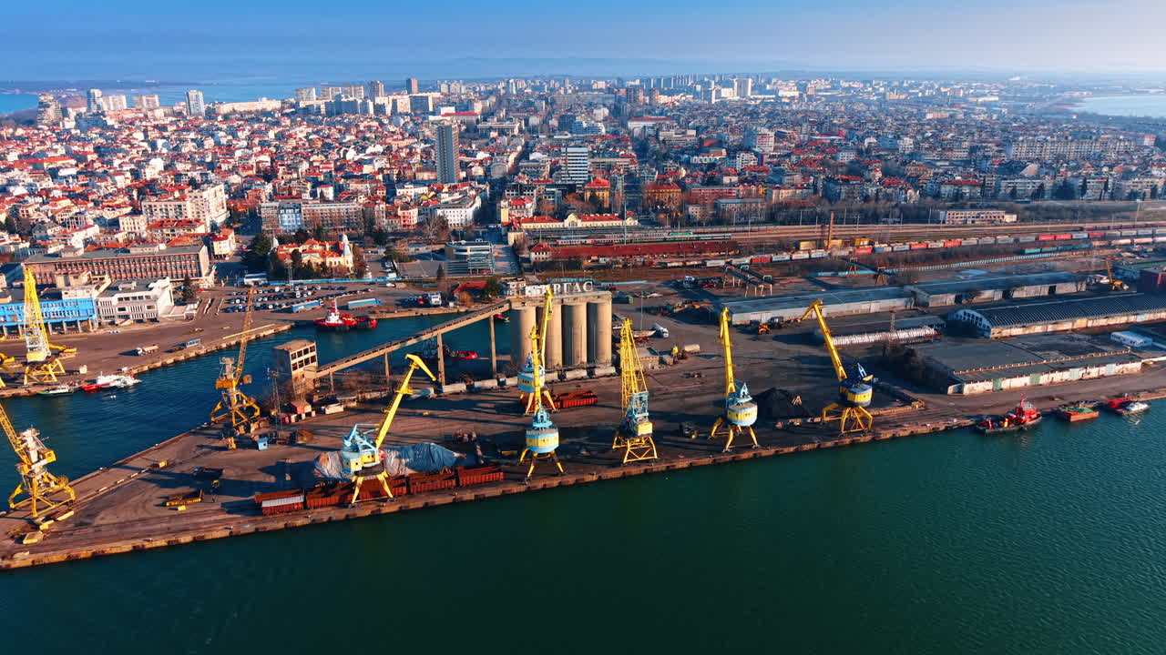 Industrial port harbor with yellow cranes and coastal city skyline. High angle view of port cranes on a pier with a dense urban city and railway in the background