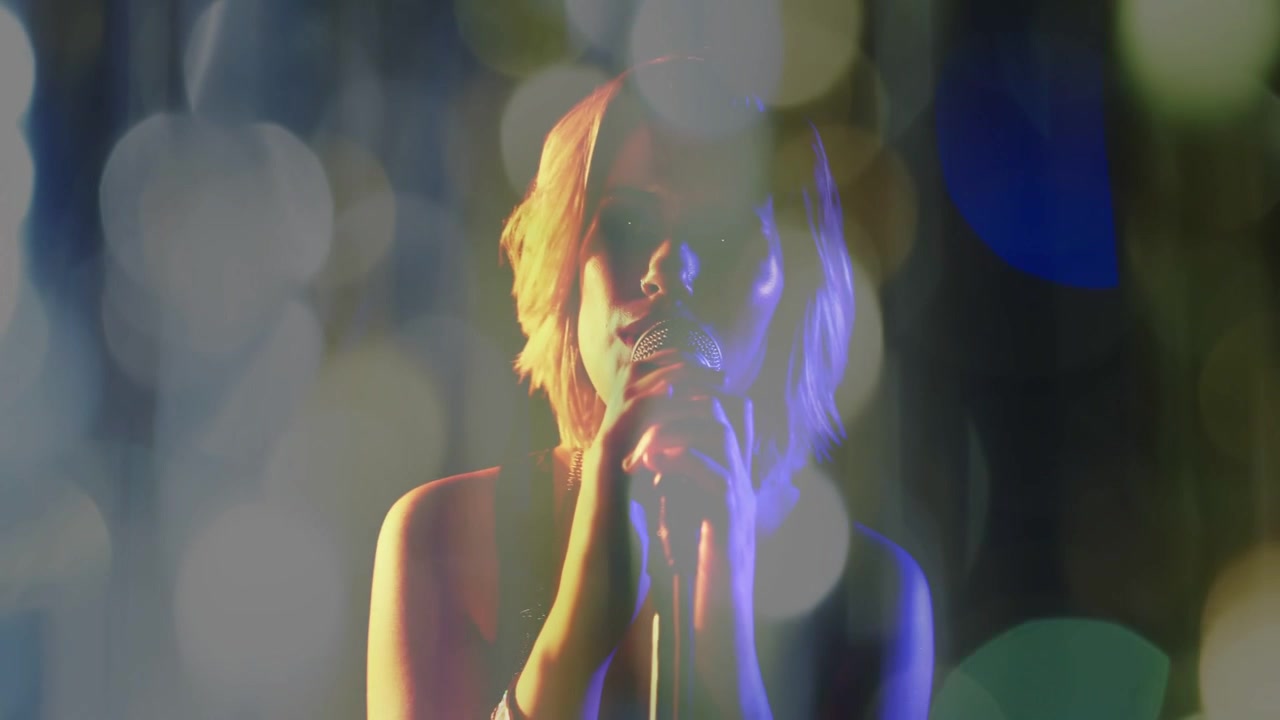 Cantante adolescente con asta per microfono, la telecamera inquadra il viso mentre canta e luci bokeh cangianti.