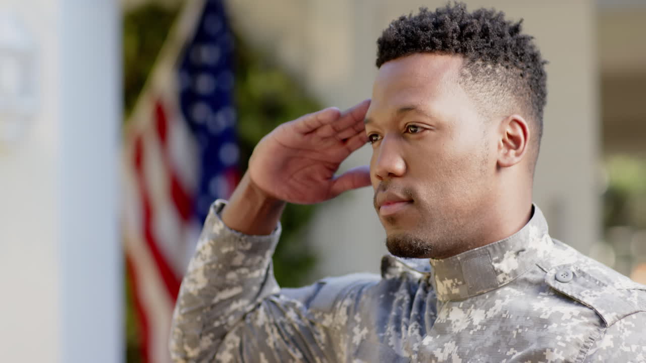 Happy african american male soldier saluting over flag of usa at home, slow motion