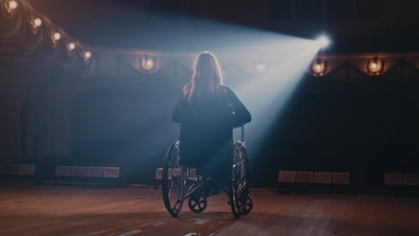 anonymous actress on wheelchair starting to play back view of anonymous woman on wheelchair riding on stage in spotlight and starting performance in theater