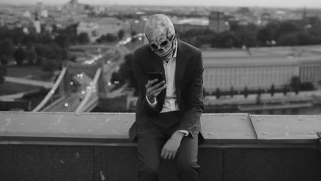 person in skull mask sitting on a rooftop using a phone in a black and white cityscape