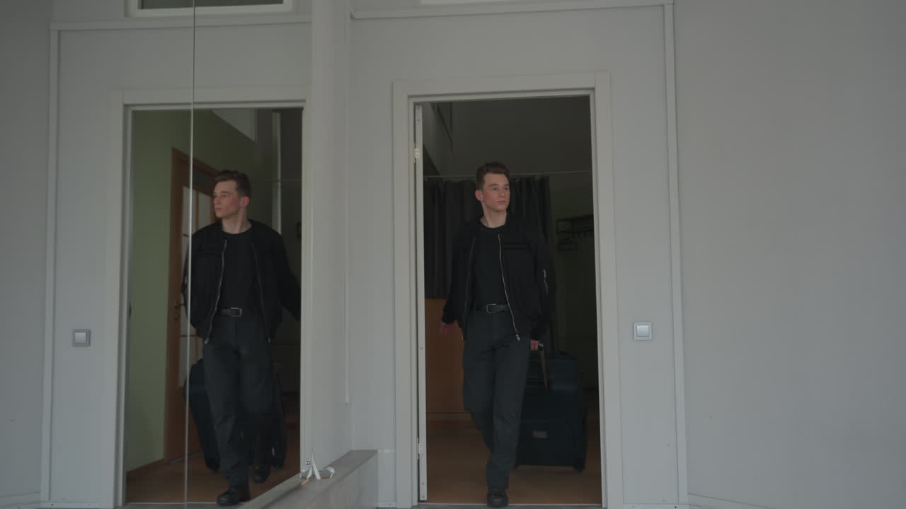 High school student walking out of room carrying travel bag leaving door open behind with large vertical mirror on wall reflecting scene interior corridor
