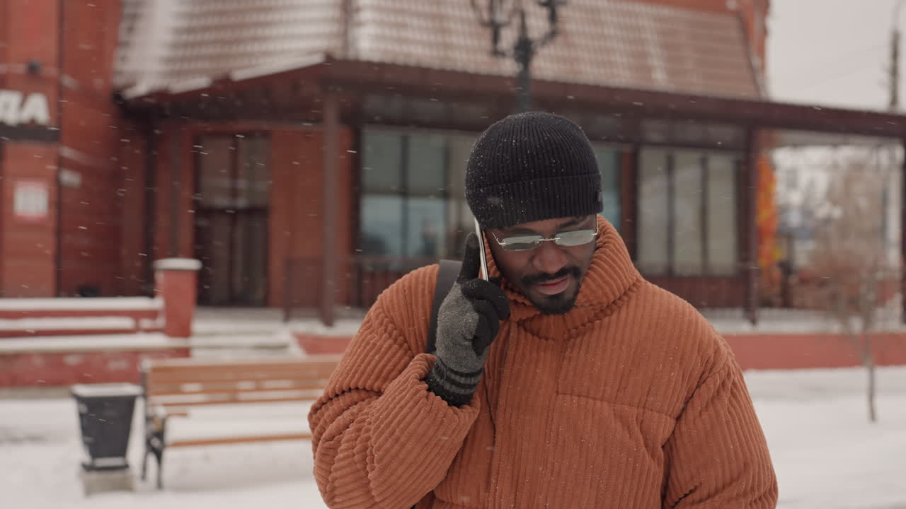 Uomo di colore al telefono per una chiamata urgente, che indossa un pesante cappotto arancione e un berretto di lana, in piedi in una piazza urbana innevata con vetrine, espressione preoccupata, che coordina un percorso di consegna e indicazioni, respiro visibile