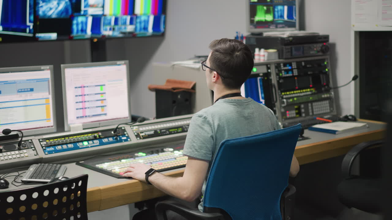 Man working in a studio, adjusting video production equipment, kneeling beside a monitor. Studio setup with bright lighting, reflective floors, and technical equipment in the background