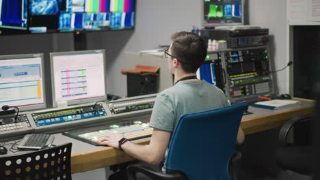 man working in a studio, adjusting video production equipment, kneeling beside a monitor. studio setup with bright lighting, reflective floors, and technical equipment in the background