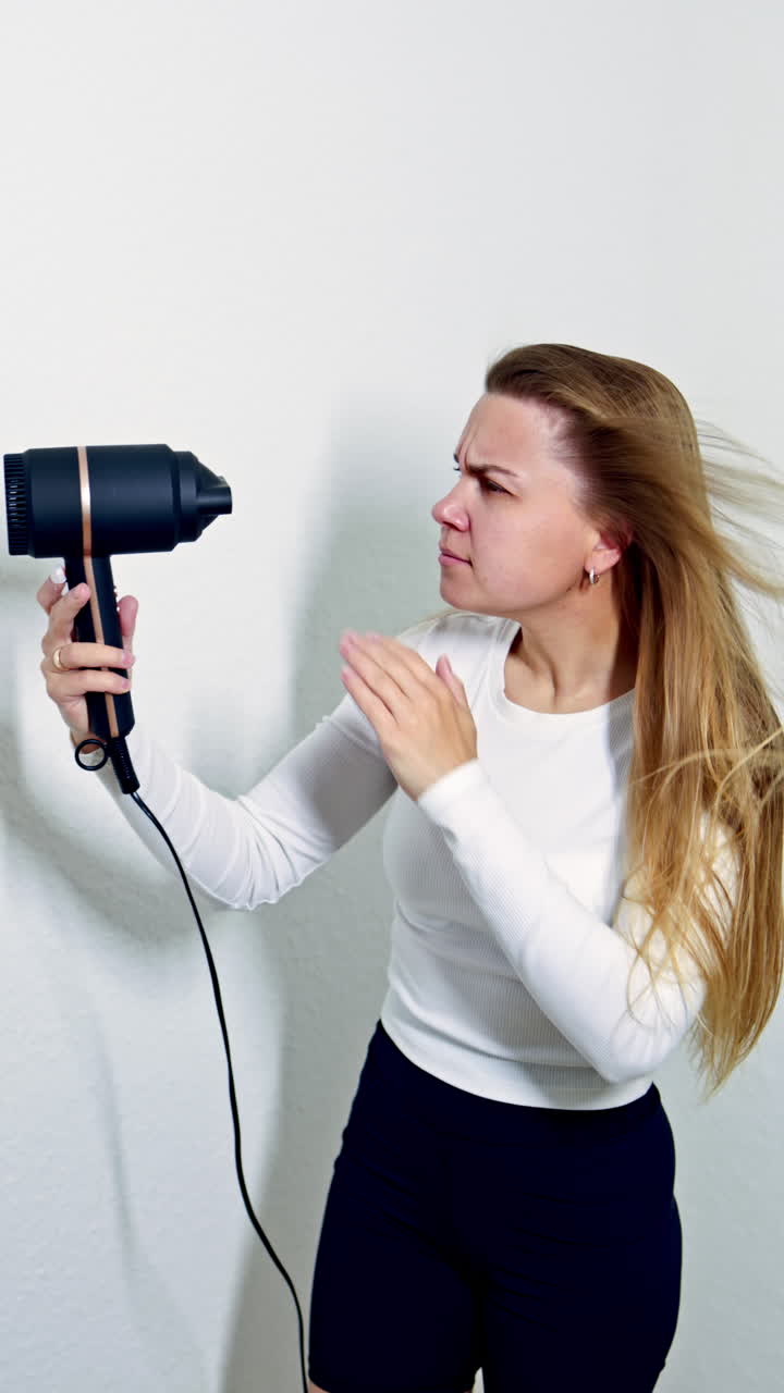 A pretty woman after washing her hair dries her hair with a hairdryer and sings. A young woman admires her hair after care procedures. Care for golden skin and long hair