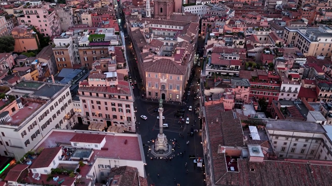 Traffico e pedoni si muovono attorno alla Colonna dell'Immacolata Concezione mentre strette strade convergono nella vivace luce serale.