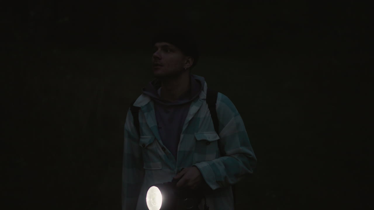 caucasian hiker at night holding flashlight wandering through dark meadow with backpack and plaid jacket, beam slicing gloom, looking up at sky, cinematic lens flare, quiet suspense and wonder