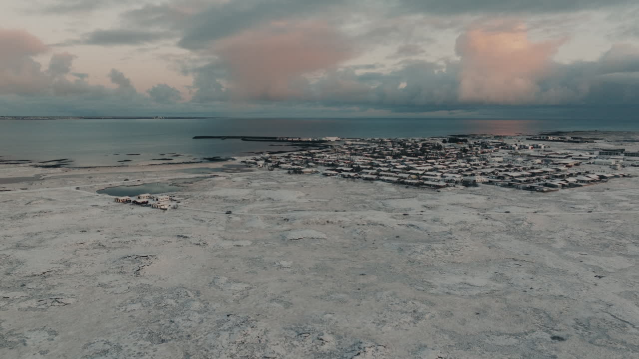Drone-ilmakuva pienestä rannikkokaupungista Islannissa lumen peitossa talvella. Syrjäinen pohjoismainen asutus jäätyneen maaston, valtameren ja dramaattisen auringonlaskutaivaan ympäröimänä.