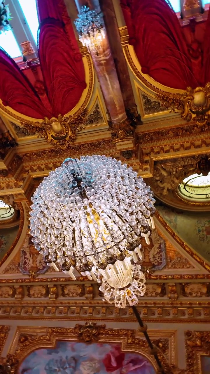 Lampadario di cristallo ornato contro un soffitto dorato con velluto rosso. Inquadratura dal basso di un grande lampadario di cristallo, modanature dorate e drappeggi rossi su un soffitto decorato