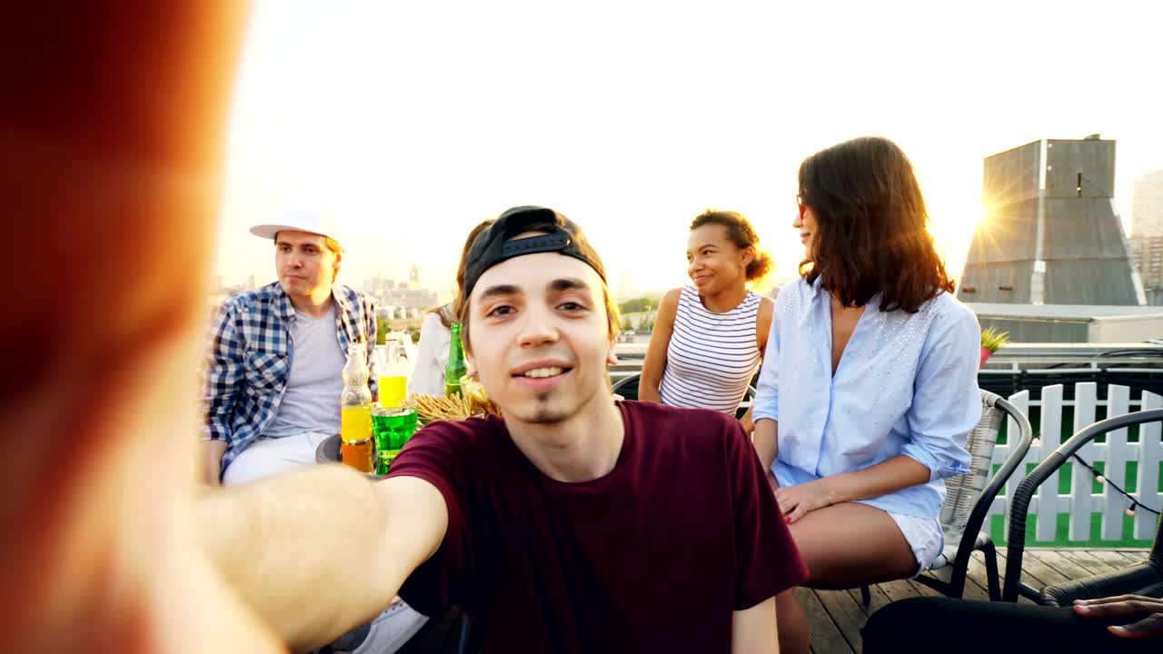 Point of view shot of young guy in cap taking selfie and posing while his friends are enjoying rooftop party and chatting sitting at table on sunny summer day.