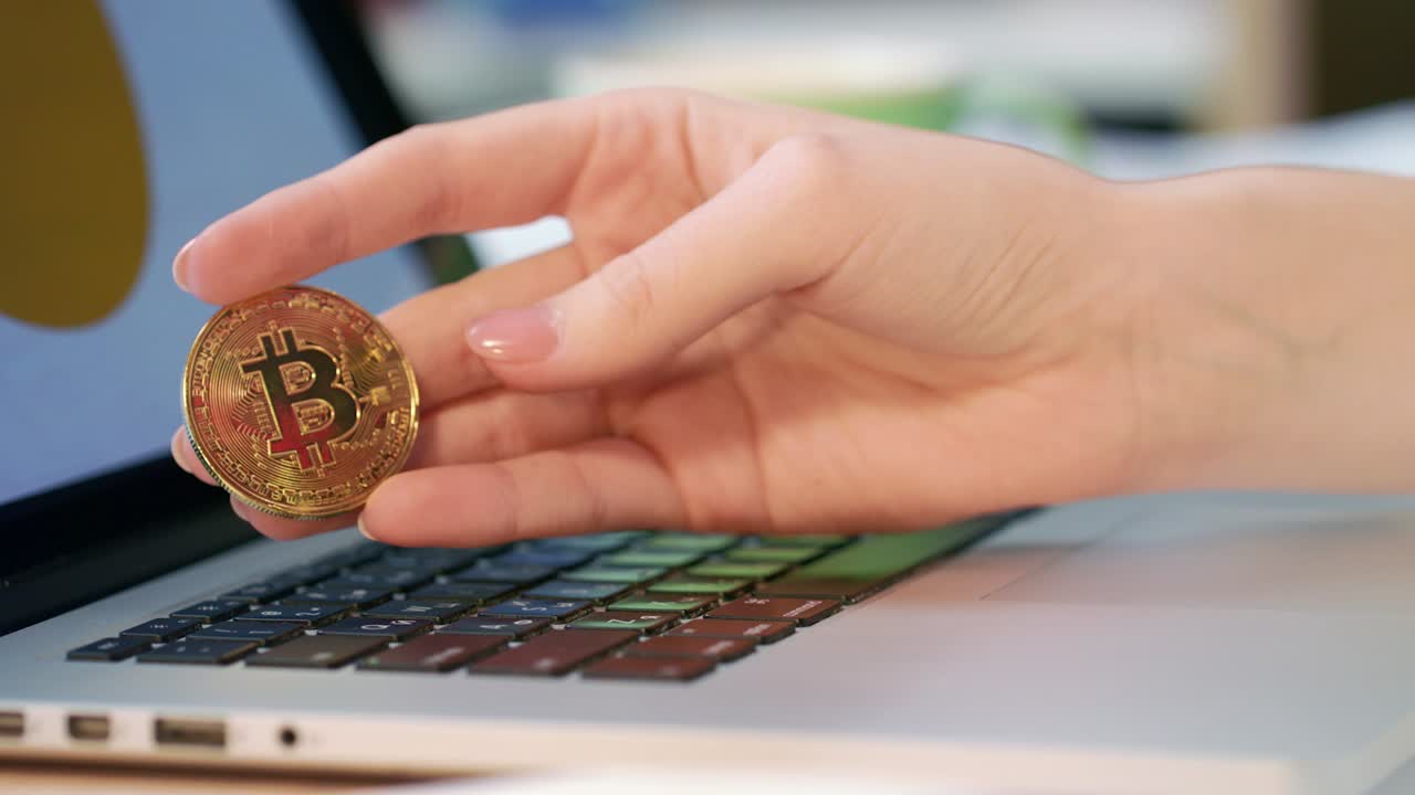 mujer con la mano sujetando una moneda de oro bitcoin cerca de un ordenador portátil