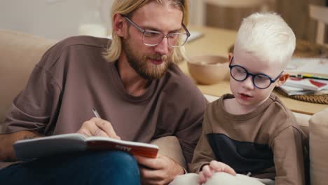 A-blond-man-in-glasses-with-a-beard-helps-his-little-son,-an-albino-boy-in-glasses-with-white-hair,-to-sort-out-his-homework-assignment-and-read-the-sentence-correctly.-Blond-man-doing-homework-with-his-little-albino-boy