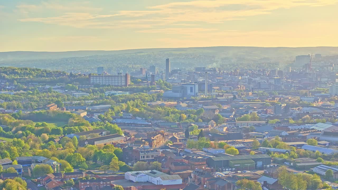 Aerial shot captures Sheffield's dense city center with high-rises, industrial rooftops, trees, and soft shadows stretching toward distant hills beyond Don Valley Bowl, slow motion