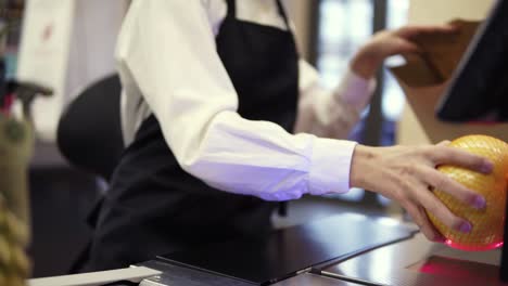 Unrecognizable-slender-saleswoman-in-white-shirt-and-black-apron-scanning-product,-fruits-at-checkout-counter-in-store-and