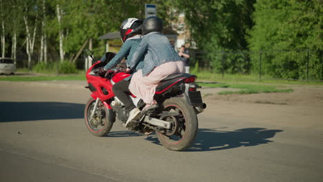 two women ride slowly on a power bike through an urban road, the passenger holds onto the rider, blurred views of people, parked cars, and tall trees are seen in the background