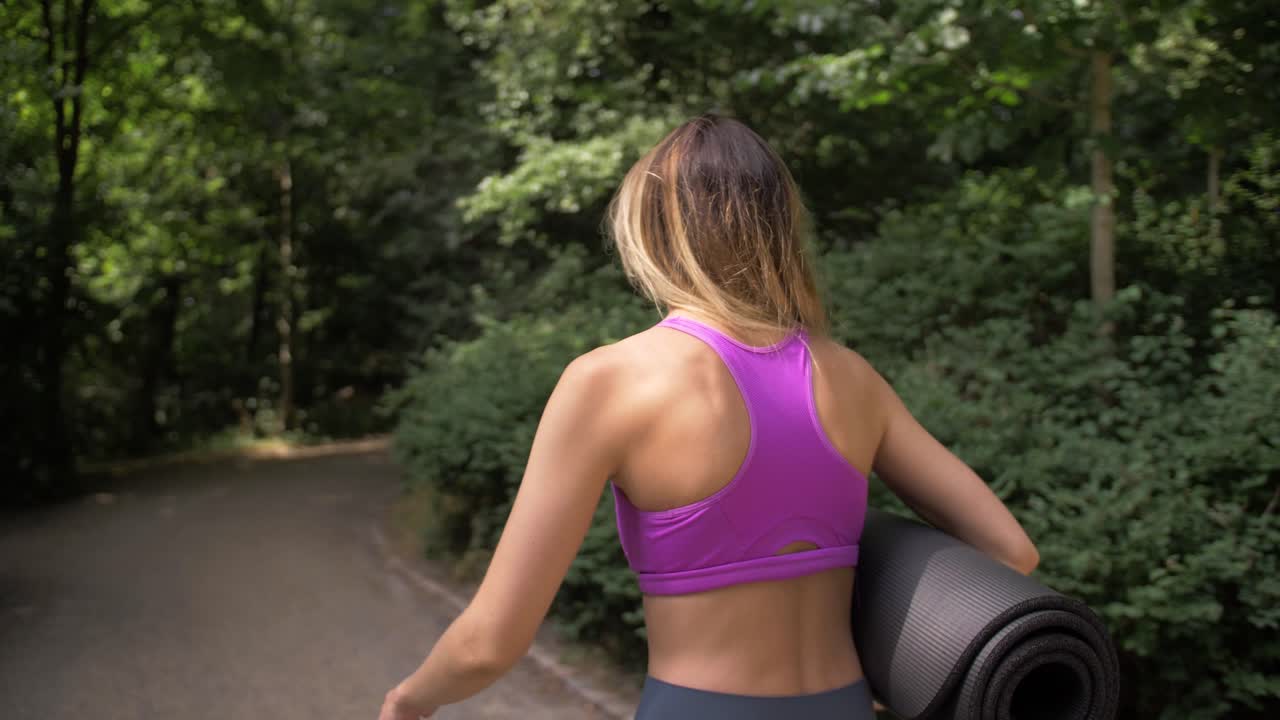 vista de cerca desde detrás de una mujer con cabello largo y rubio caminando por un sendero del parque llevando una estera de yoga
