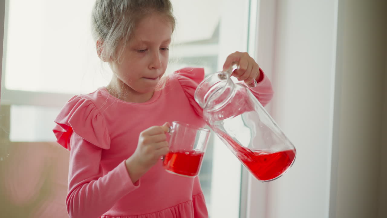 Little princess seated by sunny window handling glass jug to pour vibrant red drink into cup, then lifting cup for sip, sunlight highlighting translucent liquid and focused expression on young girl
