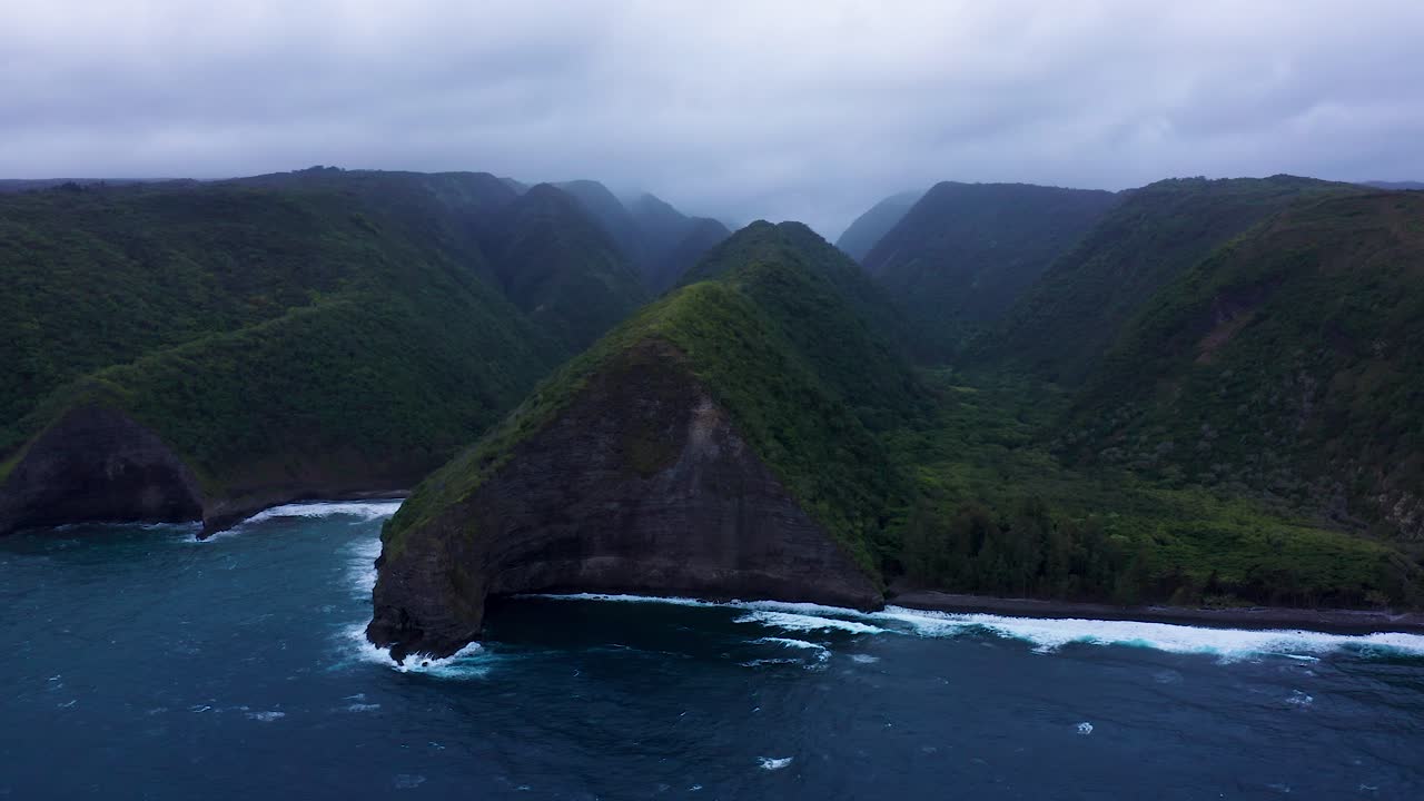 Towering emerald cliffs plunge into dark blue waves, wrapped in drifting mist and crowned by heavy clouds for a wild, untouched coastal scene.