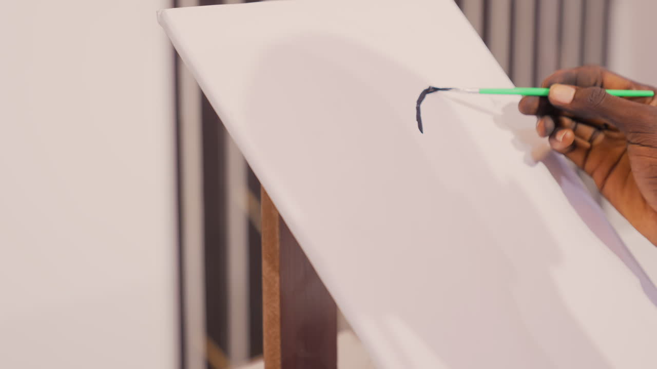 Tratto nero su tela bianca, artista che dipinge in primo piano, mano che tiene un pennello con manico verde applica pigmento fresco su un cavalletto di legno in uno studio luminoso, una ripresa ravvicinata e intima evidenzia texture, gesto e concentrazione