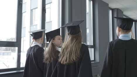 estudiantes de jardín de infantes con toga y birrete sosteniendo un diploma de graduación y caminando por el pasillo de la escuela. luego, su maestro se acerca y habla con ellos.