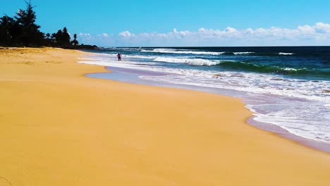 HD-Hawaii-Kauai-slow-motion-static-wide-shot-of-beach-in-lower-and-left-frame-with-a-person-standing-along-edge-of-ocean-waves-crashing-in-from-right-to-left-with-partly-cloudy-sky