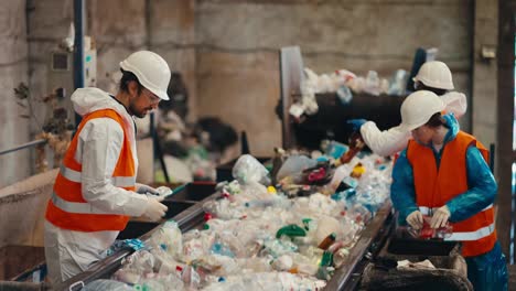 disparado desde el lado tres trabajadores en uniformes blancos en chalecos naranjas clasifican la basura cerca de una cinta transportadora transfiriendo botellas de plástico por color a las cajas apropiadas en una planta de reciclaje de residuos