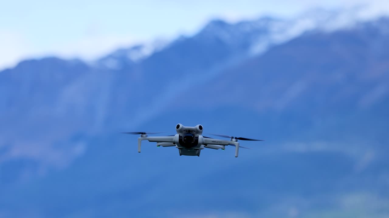 Quadcopter drone approaches camera with steady movement, daylight, distant snow-capped mountains, wide shot