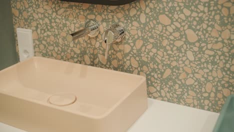 a detail shot of a chic bathroom corner, with a peach-colored basin set against a terrazzo-tiled wall and polished chrome faucets, conveying a modern and artistic flair