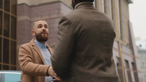 caucasian businessman talking on the phone and looking at the open notebook in his hands in the street in autumn, then an african american man approachs to him and shake hands