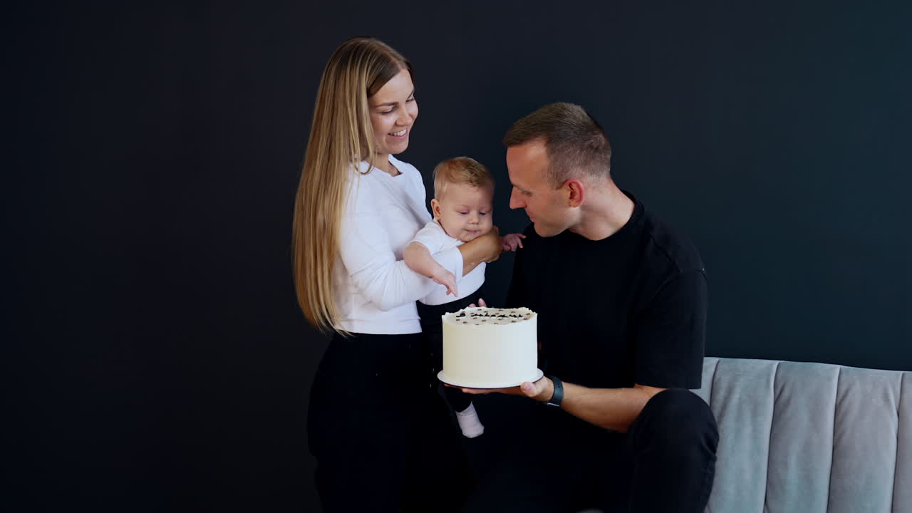 Man sitting on the sofa holding a white cake. Woman holding a baby near the cake. Black backdrop.