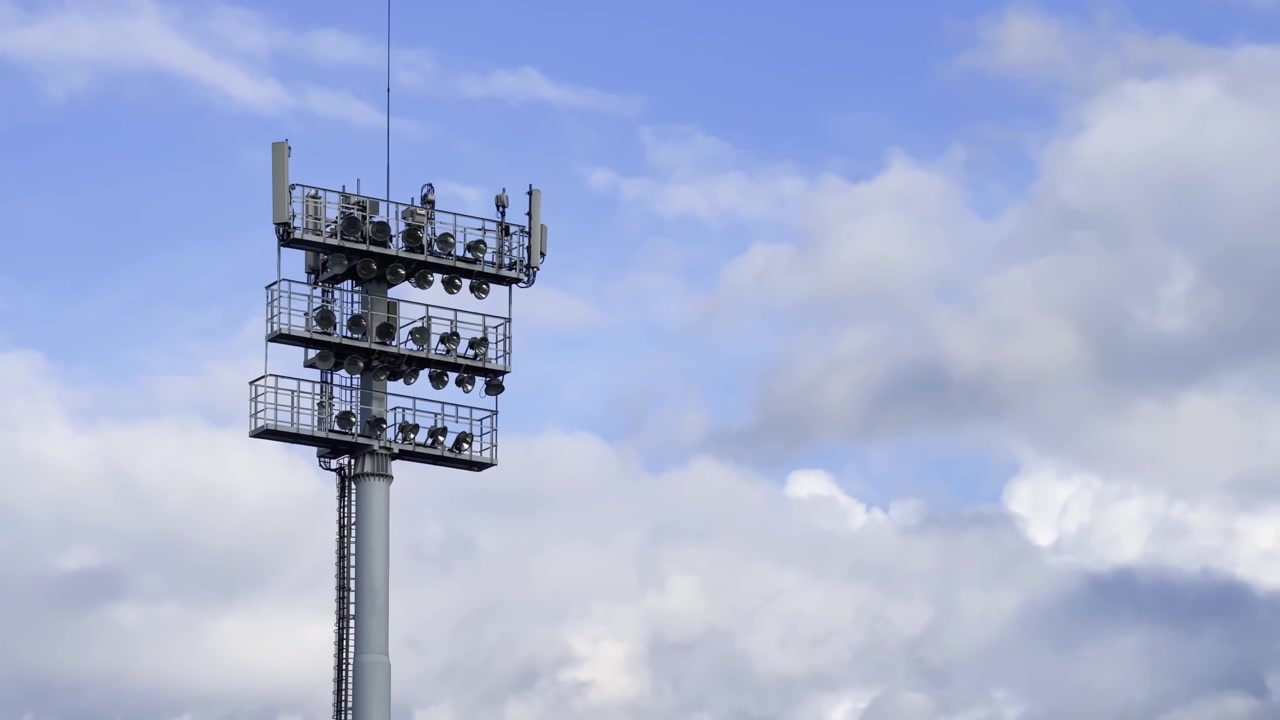 Torre faro dello stadio con proiettori e antenne per telecomunicazioni contro un cielo azzurro nuvoloso, che mostra l'infrastruttura di un impianto sportivo di giorno.