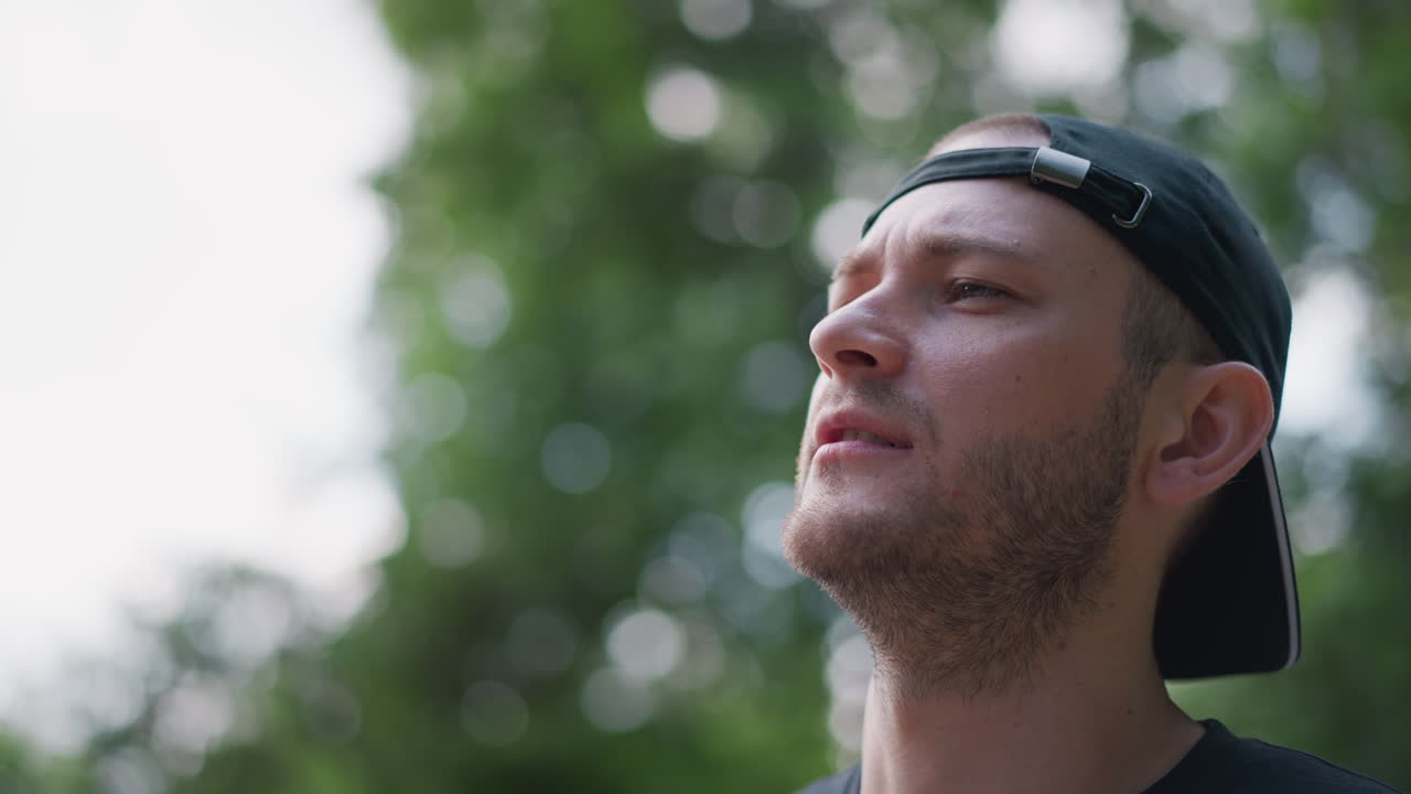 Caucasian Man Drinking Water From Bottle Outdoors, Wearing Backward Cap And Light Stubble, Park Foliage Blurred In Background, Daylight Bokeh, Closeup Profile Shot Conveying Refreshment