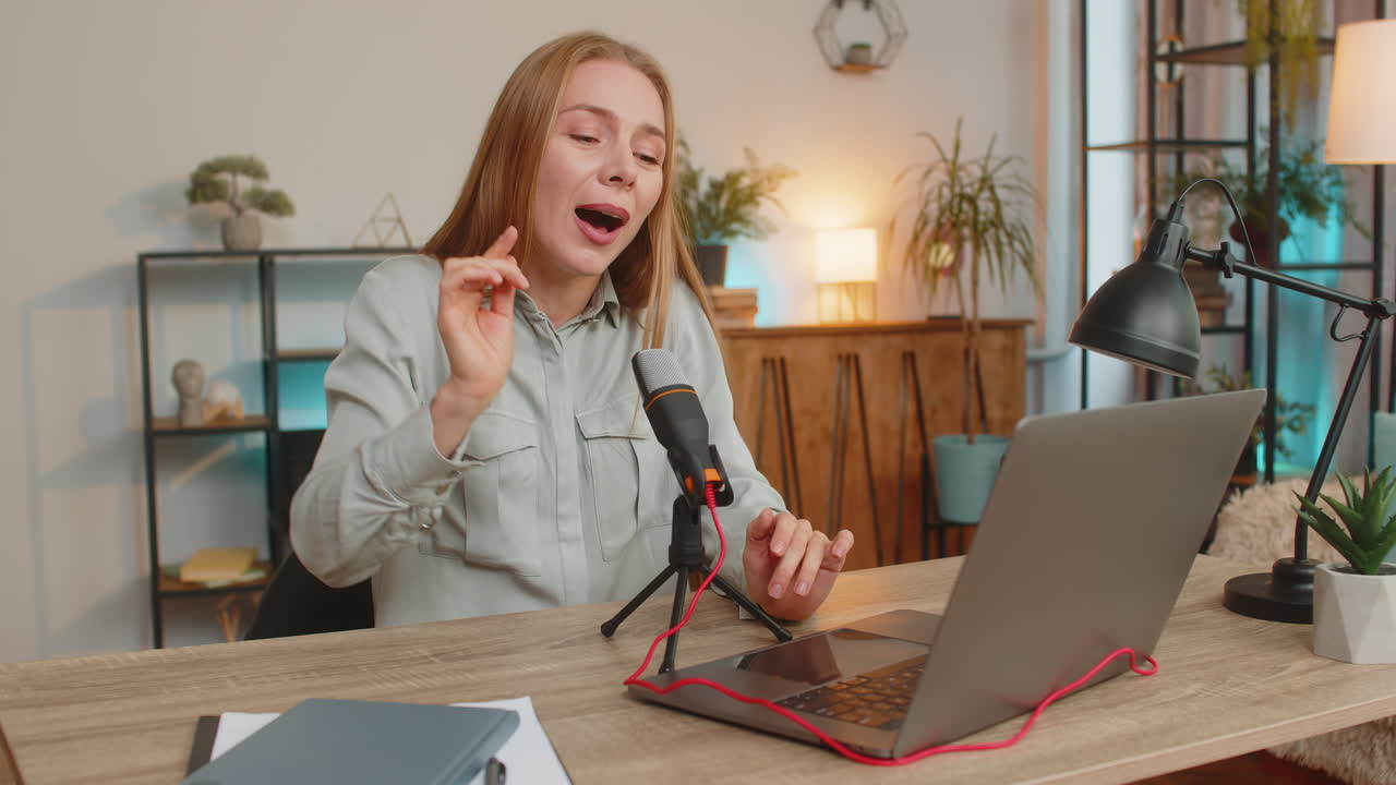 Mature positive caucasian woman blogger singer using laptop while singing into microphone at table