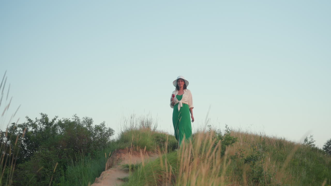 Woman in green dress and white hat walking down grassy hill holding wine glass under clear blue sky, surrounded by wild nature, enjoying peaceful outdoor moment during golden hour