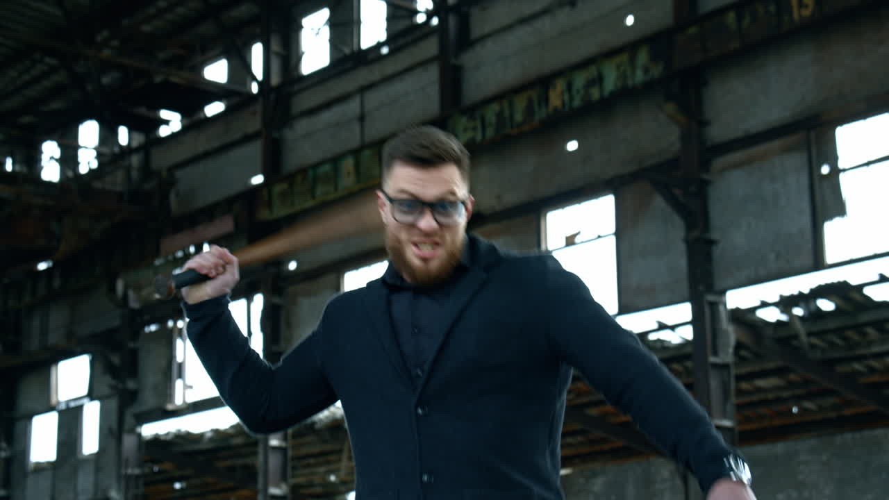 Strong bearded young man with bat. Guy waves a baseball bat on the background of abandoned house. Bandit looks at camera and shouts.