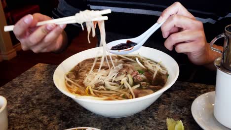 adult male asian man hand stirring pho vietnamese soup with long noodles pork and some vegetables holding a chinese soup spoon with hoisin sauce mixed with spicy hot sriracha sauce with chop sticks