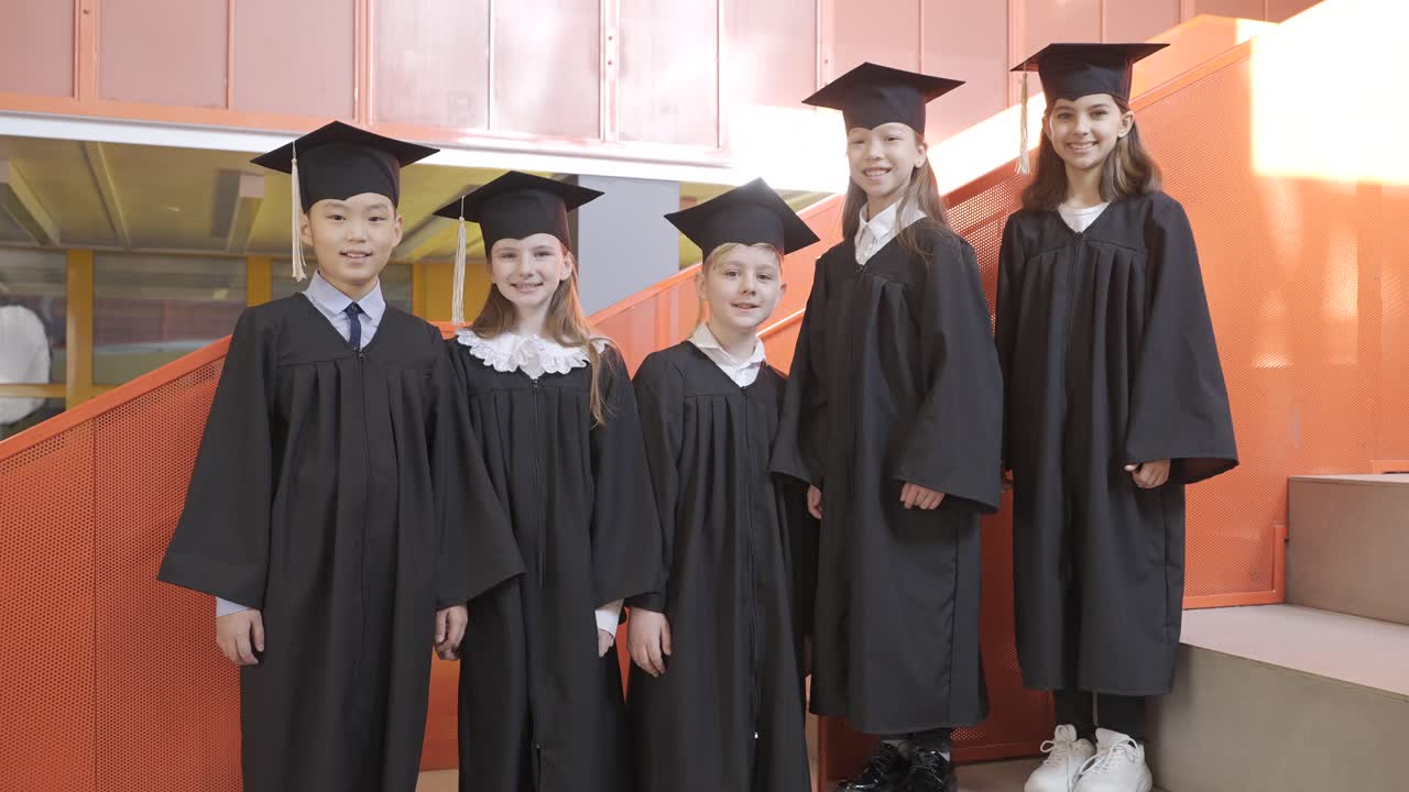 유치원 졸업식에서 카메라를 보고 엄지손가락을 치켜드는 모자와 가운을 입은 행복한 유치원생들의 확대