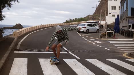 Man-longboarding-on-a-road-close-to-coastline-in-cloudy-weather.-Tall-man-in-hoodie-practicing-skateboarding-by-high-angle-road.-Slow-motion