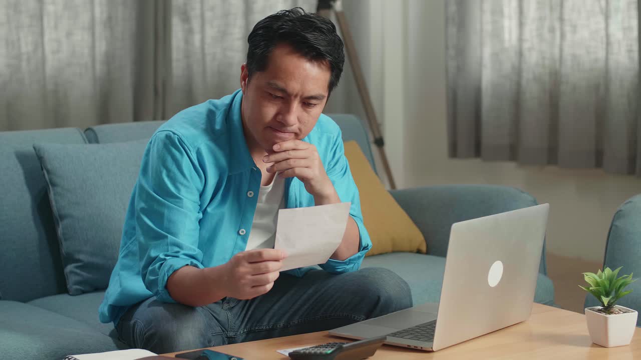 아시아인 남자가 집에서 노트북으로 일하기 전에 청구서를 보고 생각하고 있습니다.
