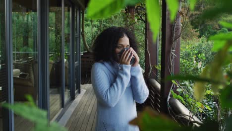 Happy-mixed-race-woman-enjoying-drinking-coffee-standing-on-balcony-in-the-countryside