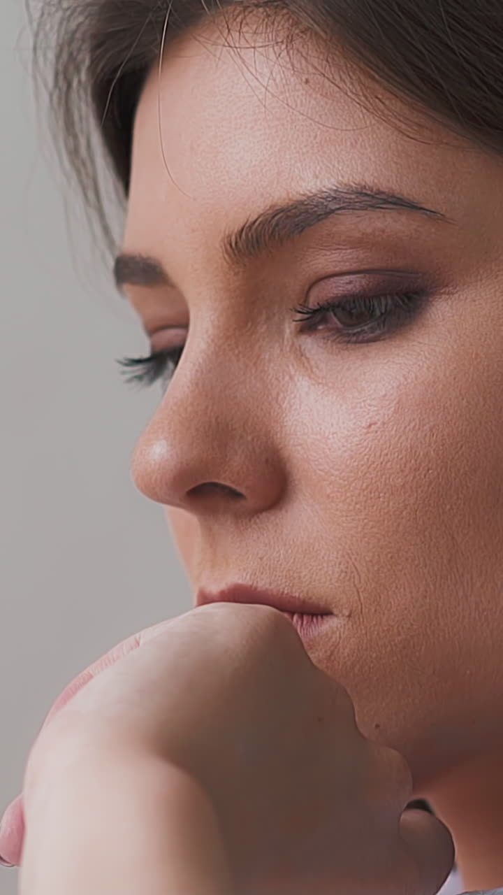Pretty unhappy young woman with dark hair touches chin by hand posing for camera on light background in room close side view slow motion
