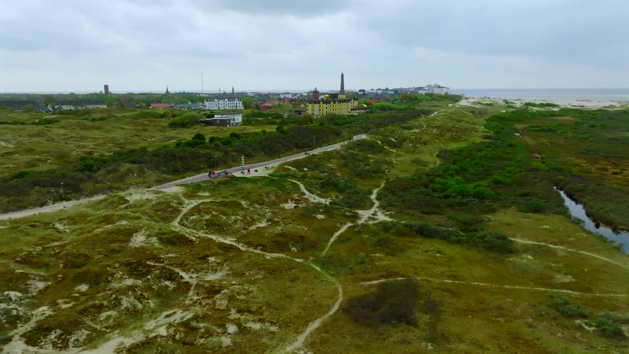 Low altitude aerial shot orbiting a milestone marking the northernmost point of Germany. Grassy dunes and green marshland with sandy trails leading to Borkum town in the distance.